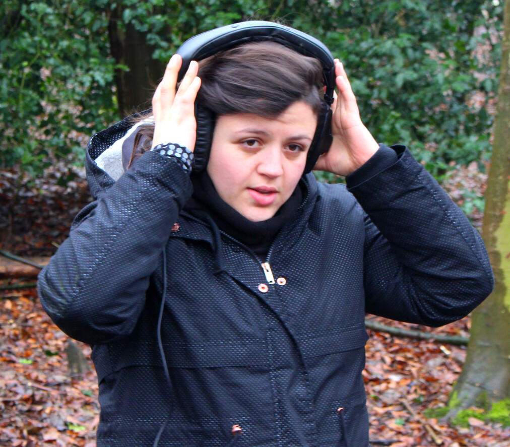 A photo of Clau in the woods. They have short dark hair, a black coat and big headphones.